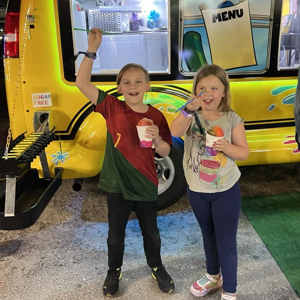 Two kids smiling and holding up their shaved ice in front of the bus