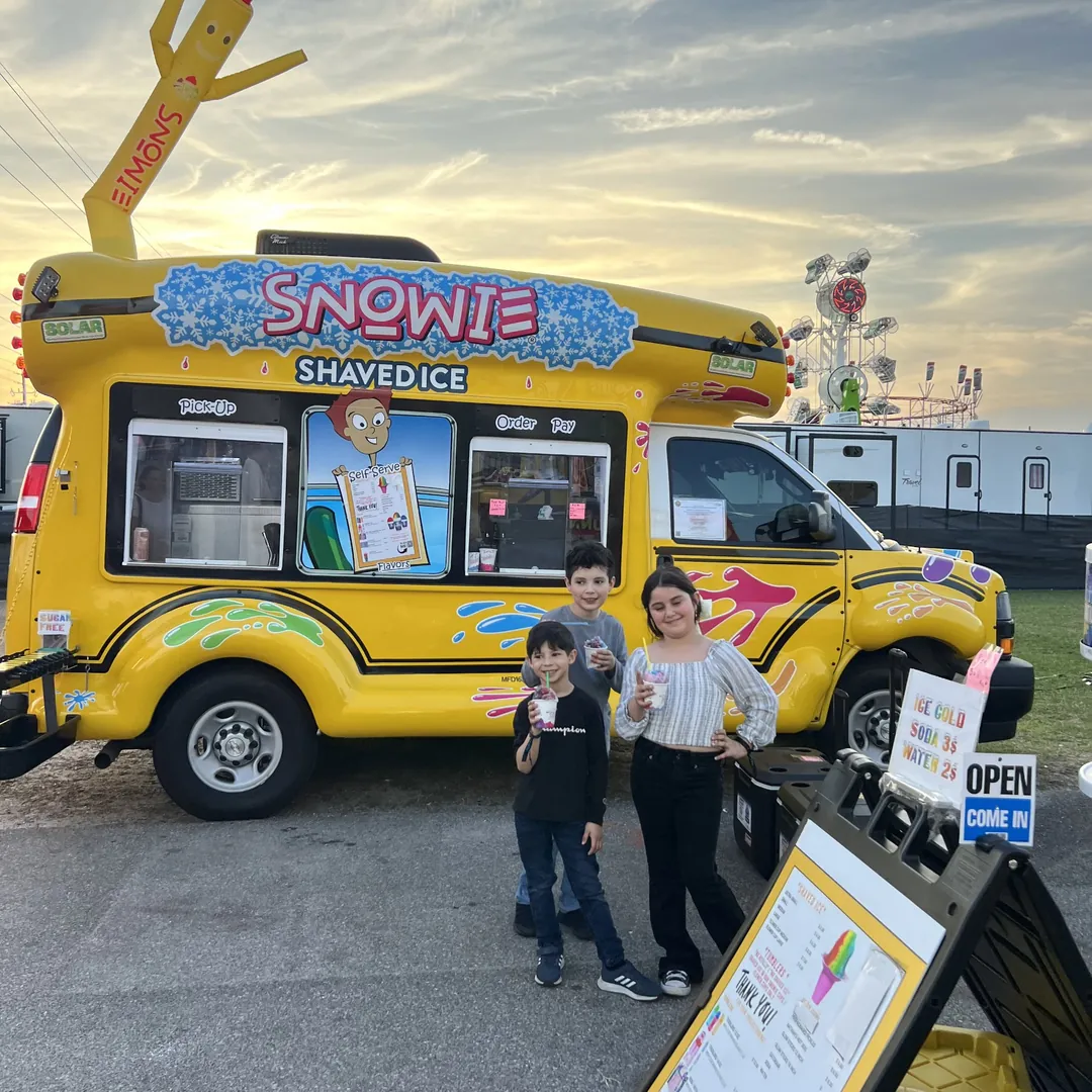 Three kids with shaved ice in front of the bus at a fair at sunset