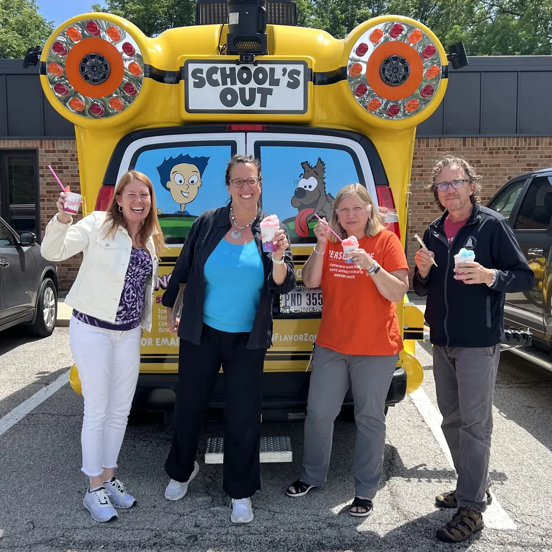 Four adults holding up shaved ice at the back of the bus, under its School's Out sign
