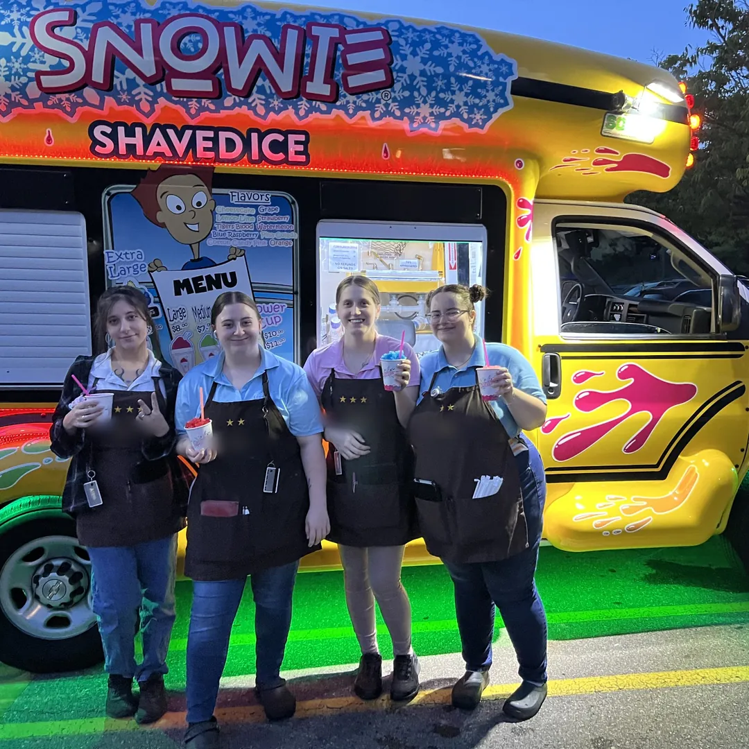 Four servers in aprons with shaved ice in front of the lit-up bus at dusk